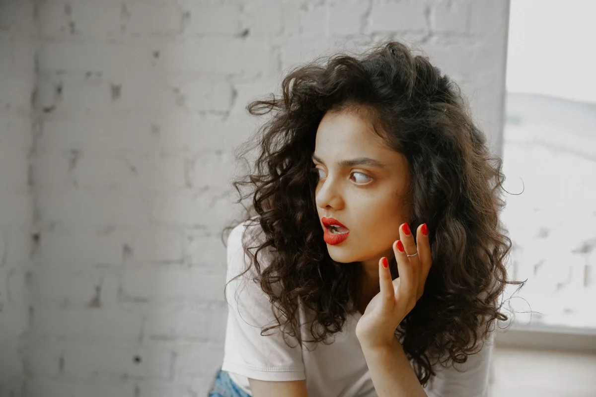A woman's shocked expression captured in profile, hands raised in distress, against a blurred interior background with warm lighting