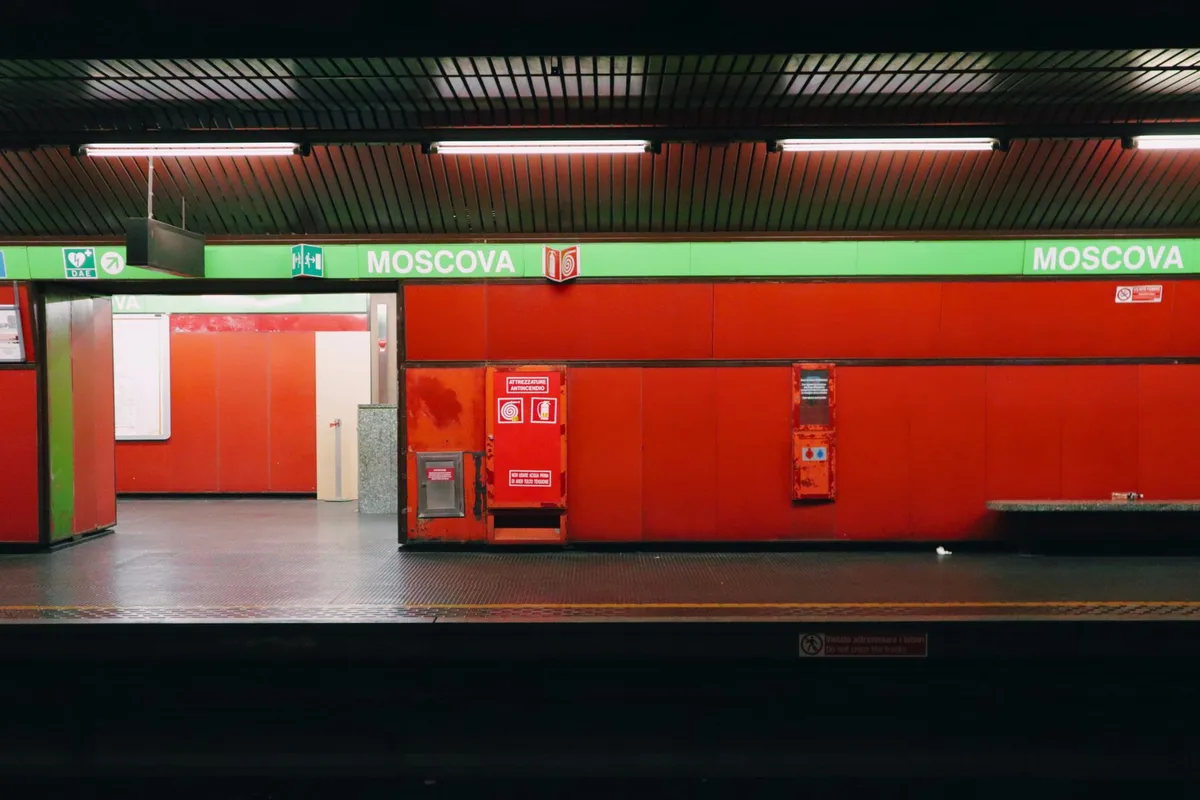 Two figures standing in a modern underground transit station, separated by commuters, their eyes meeting across the platform with a spark of recognition, neon light reflecting on tiled walls