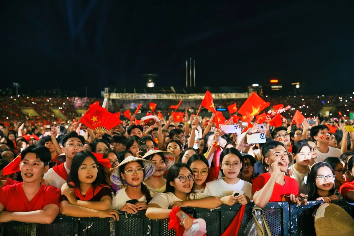 Anonymous fans surrounding a woman in white leather and cowgirl hat at a rodeo arena, arms raised in greeting, faces blurred, floodlit evening atmosphere