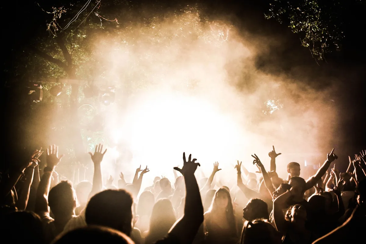 Crowd of spectators in festival attire watching a stage performance, hands raised, excited expressions, colorful clothing and hats, festival atmosphere with warm lighting