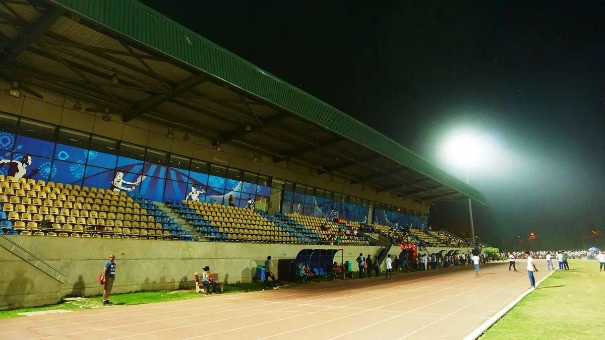A soccer stadium interior at dusk with floodlights illuminating an empty field, white field markings visible, seats casting shadows in the stands, anticipation in the air
