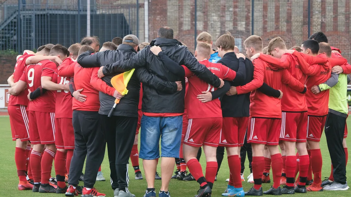 Soccer players in red and white jerseys gathered in a huddle on the pitch, heads together in focus, stadium lights in the background, determination visible in body language