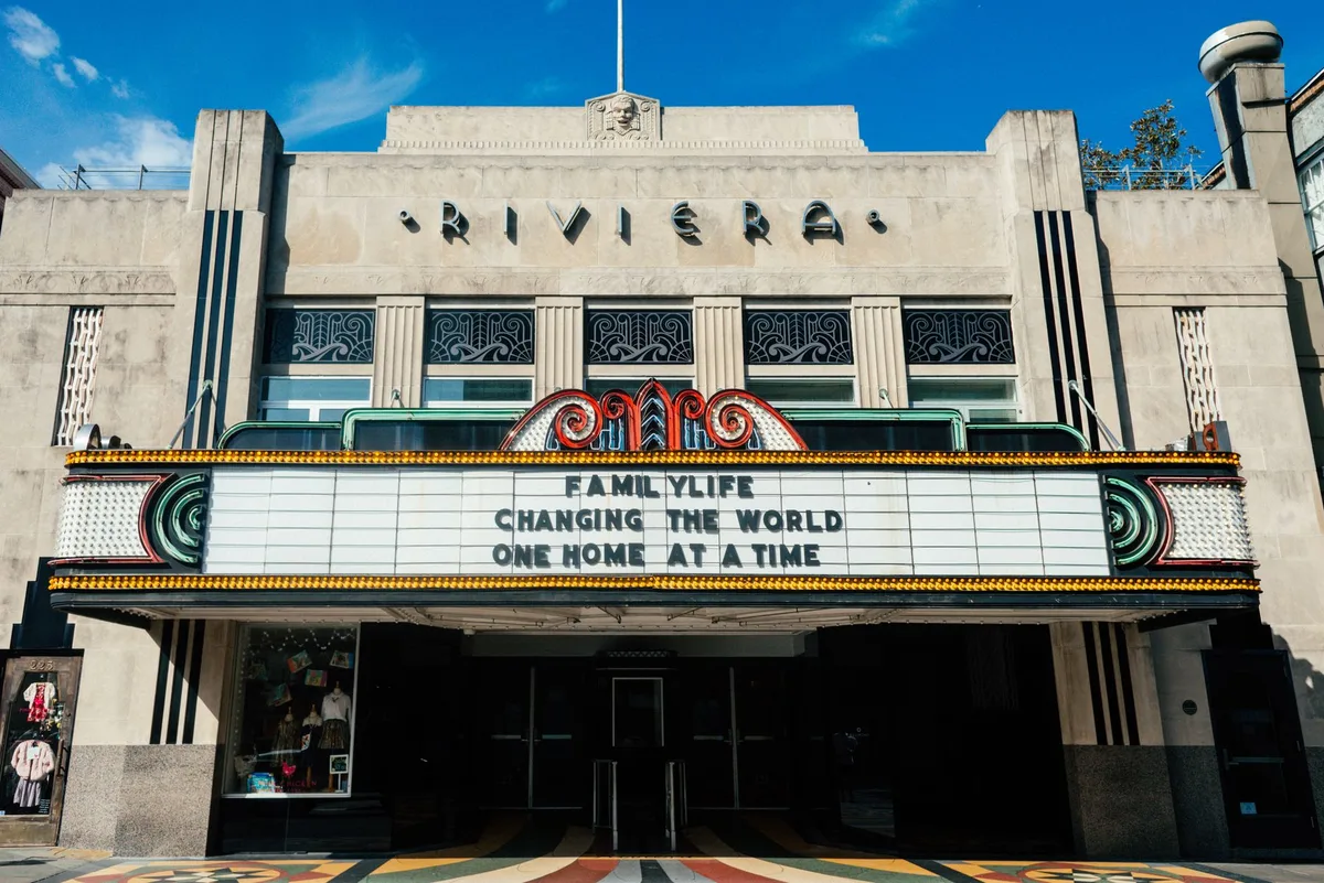 A movie theater marquee with bright lights, audience silhouettes entering through glass doors, evening glow, popcorn stand visible, anticipation in the scene