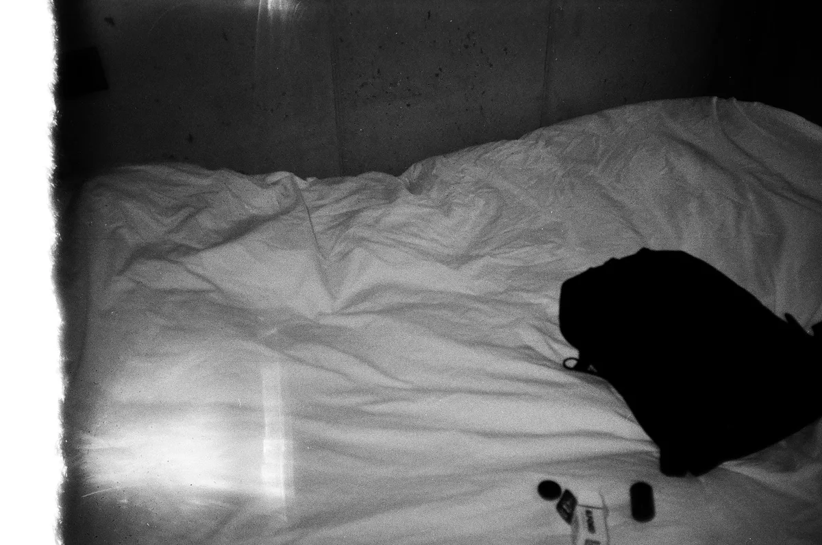 A dimly lit bedroom with an empty chair beside an unmade bed, soft morning light filtering through curtains, a family photograph on the nightstand, the room quiet and still, evoking absence and loss