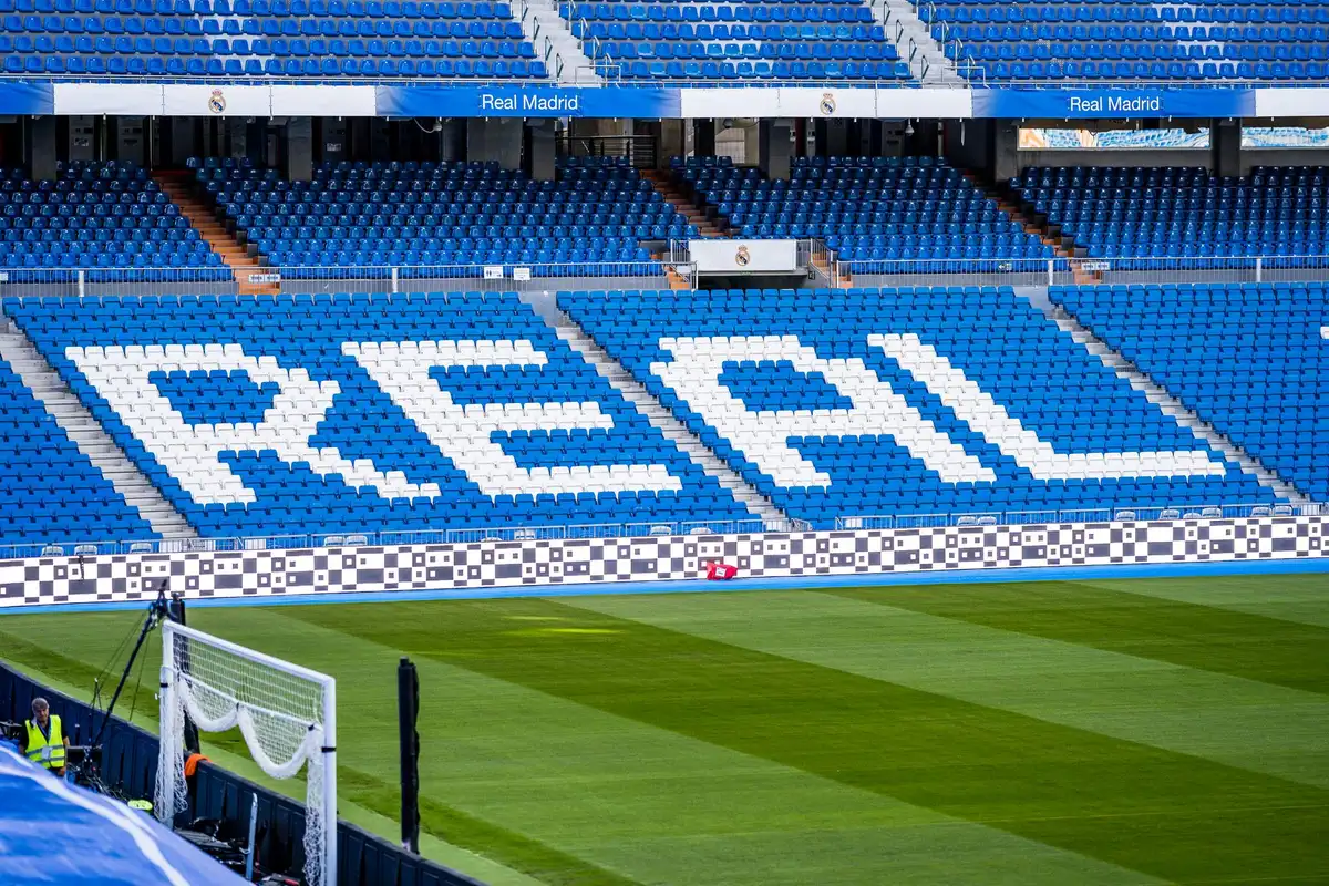 A stadium training ground with modern facilities, a young player in training gear working with the ball, the Real Madrid crest visible in the background.