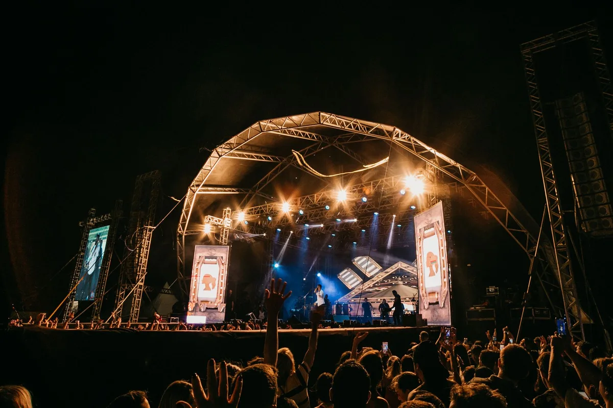 A concert stage with colorful stage lights and silhouettes of musicians performing, instruments catching the glow, a packed crowd with arms raised in celebration