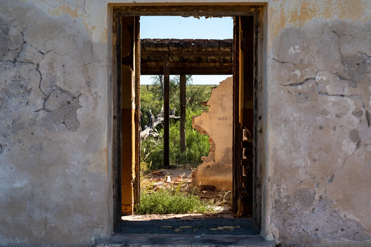 A woman in her 50s standing in the doorway of a dilapidated motel building at sunset, warm light casting long shadows across weathered walls, a knowing smile on her face, the structure behind her crumbling but full of potential.