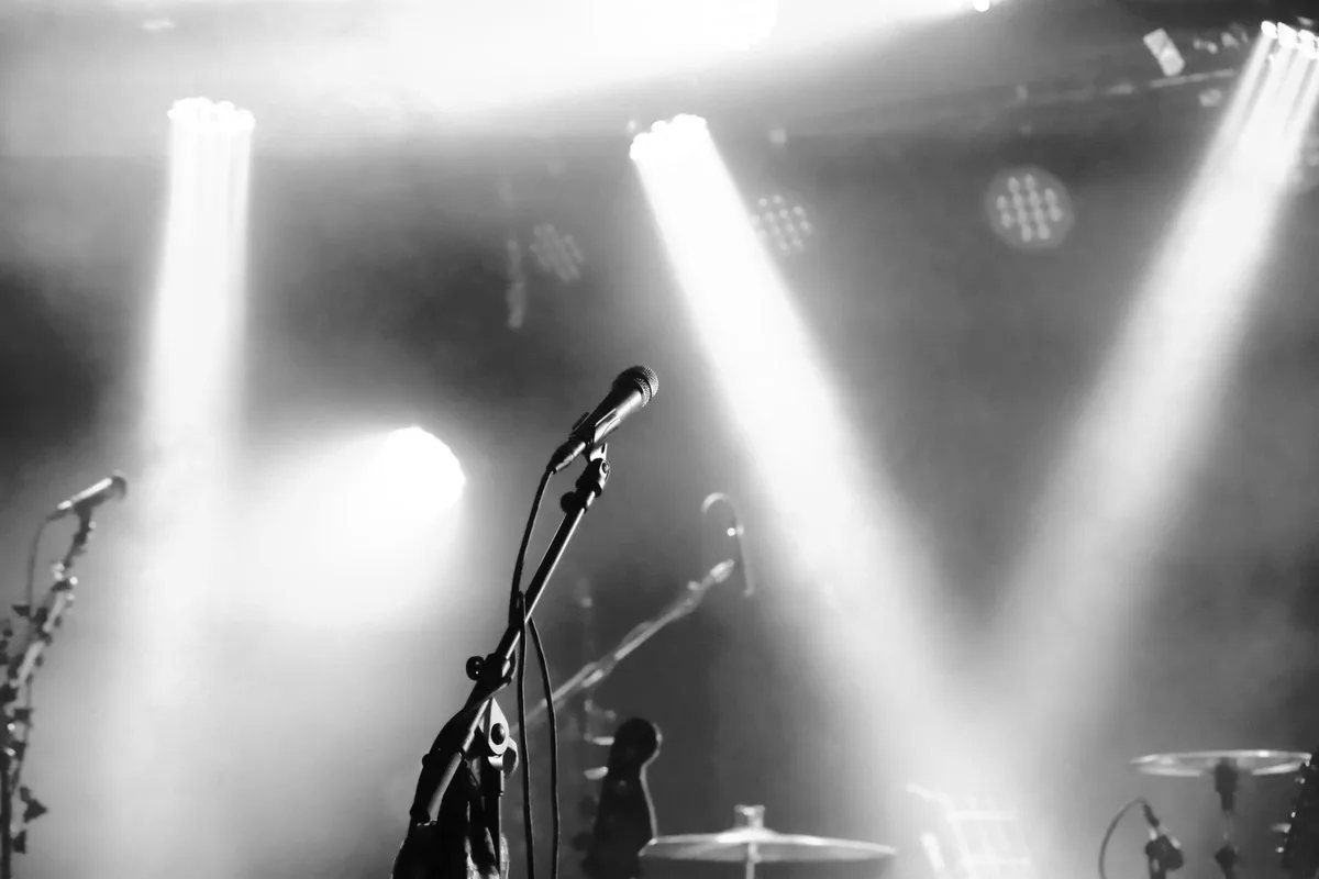 A glowing microphone on a dimly lit stage, soft spotlight illuminating the stand, a coiled cable trailing into shadow, dust particles drifting in the beam, empty concert hall seats blurred in background