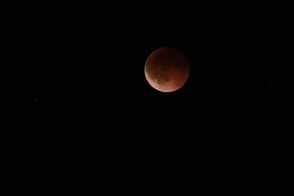 A deep partial lunar eclipse with most of the Moon covered in Earth's shadow, displaying a reddish hue in the darkest region, stars visible around it