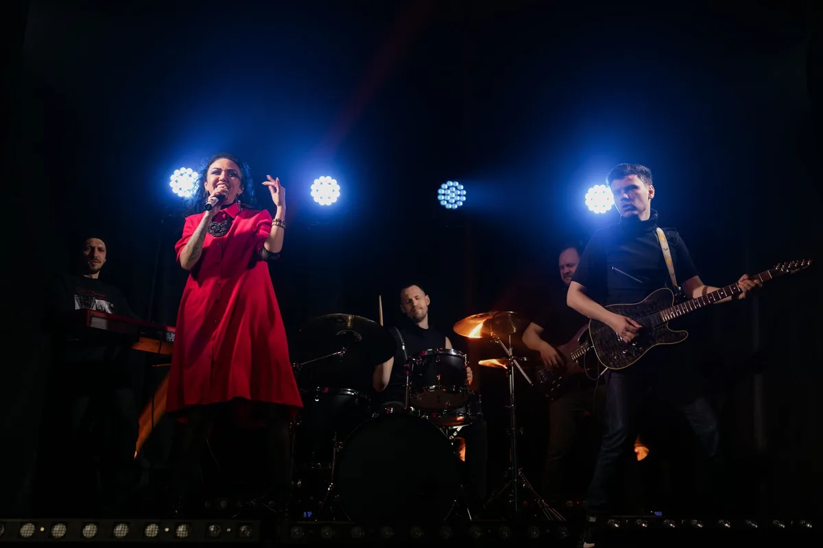 Two music mentors stand face-to-face on a brightly lit stage, evaluating talented singers in separate performance areas, one marked with vibrant pop aesthetic, the other with traditional pagode style decorations