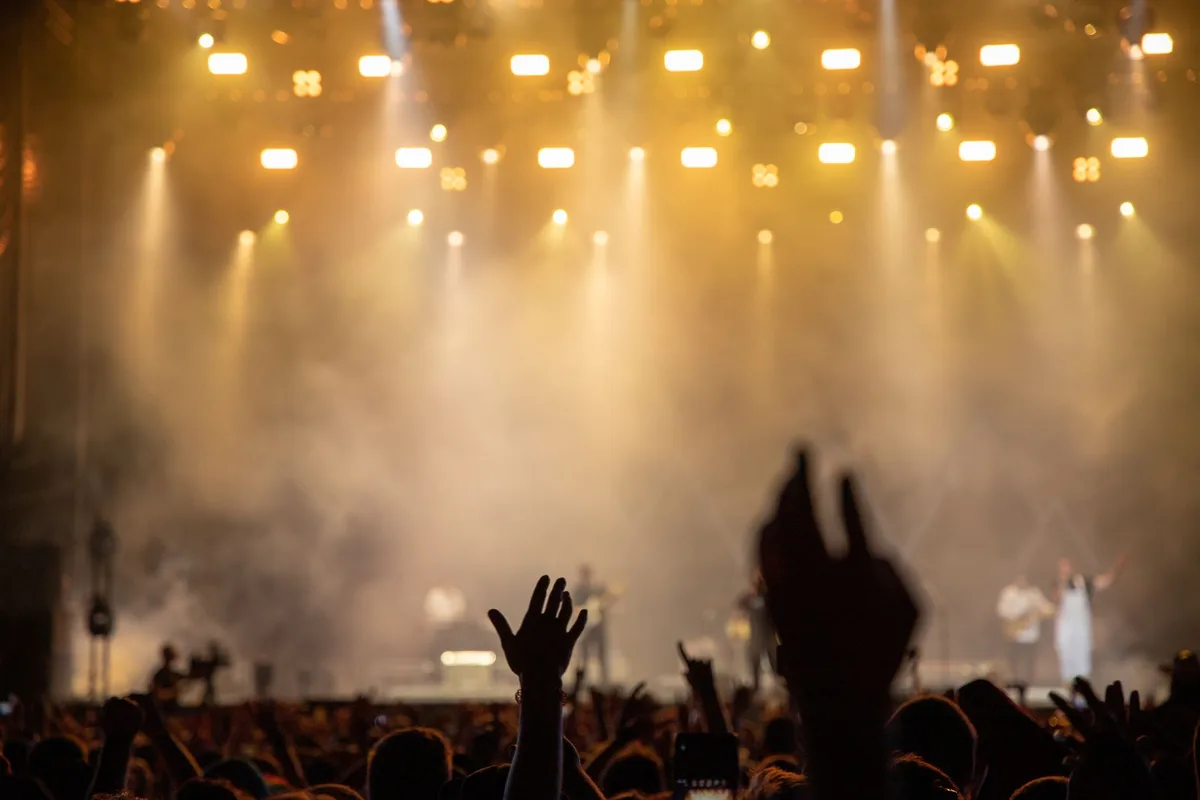 A stage filled with colorful stage lights and a microphone stand center-frame, with silhouettes of audience members in the background, representing the live performance element of the competition
