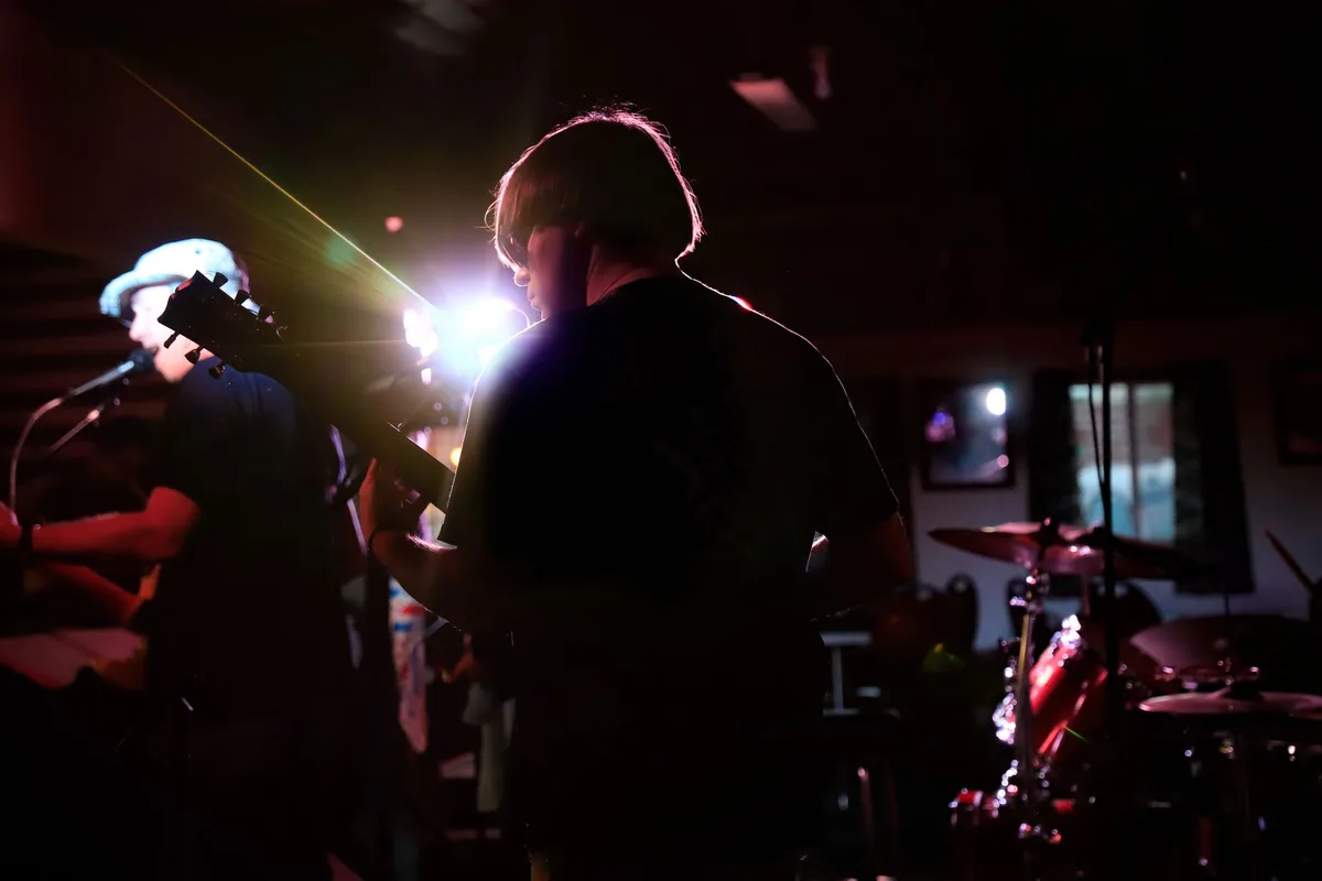 Three silhouetted figures on a concert stage under blue stage lighting, with the center figure at a drum kit, the scene capturing the iconic band formation from decades of performances.