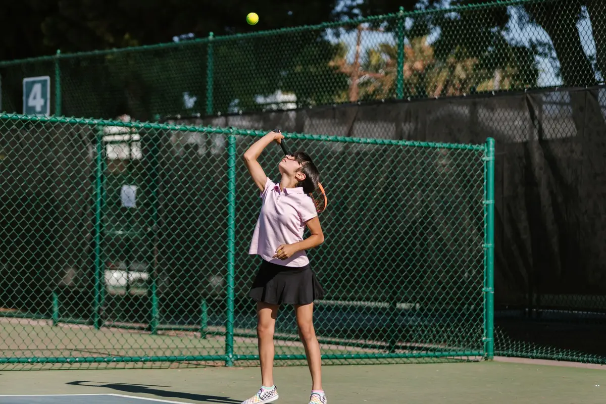 Tennis player in serving motion on a hard court, arm extended high, focused expression, stadium crowd blurred in background, bright sunlight casting sharp shadows