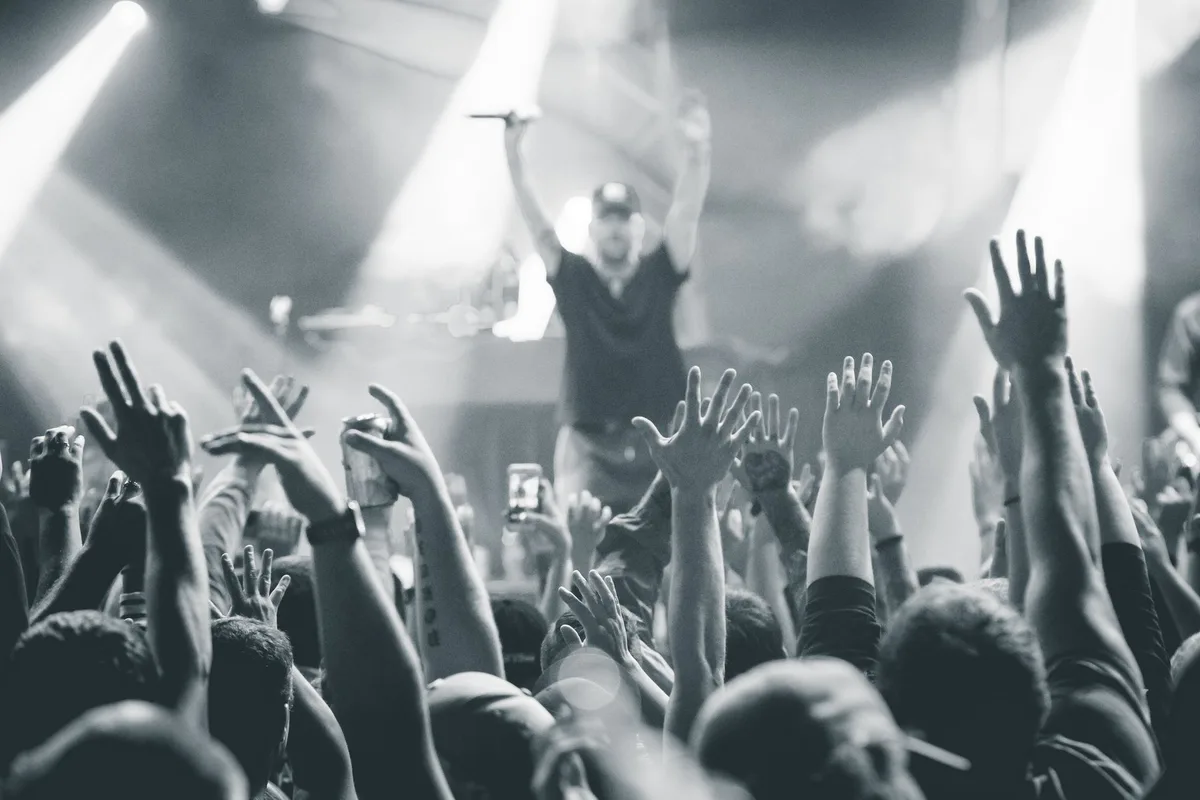 An arena stage under bright floodlights, a crowd of anonymous silhouettes with arms raised, the stage glowing with stage lights, the energy of thousands of spectators in motion, a single spotlight illuminating the performance space