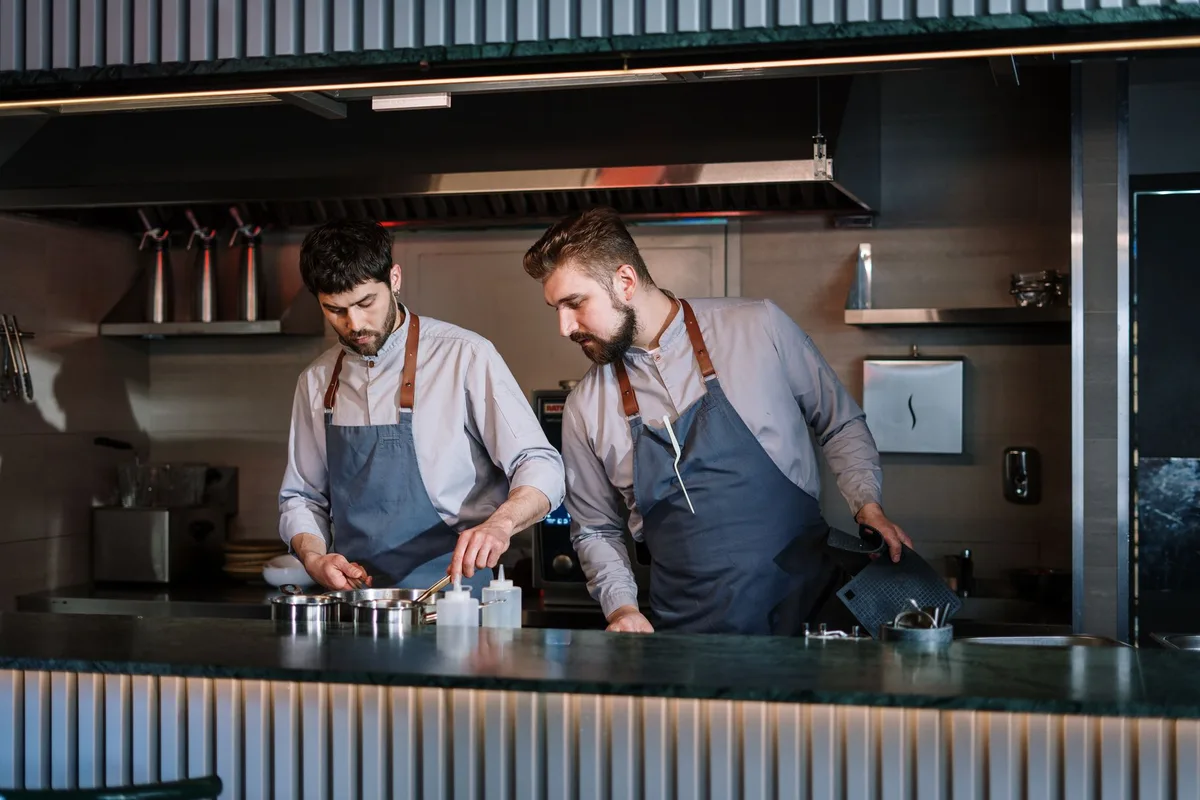 A bustling kitchen scene with three anonymous chefs in chef whites evaluating a plated dish, their faces partially obscured, focused on examining the food, professional kitchen equipment visible in soft focus behind them