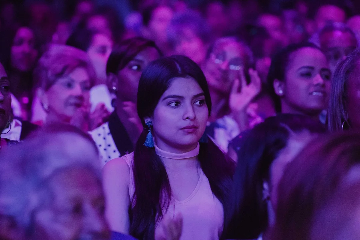 An intimate moment of a young presenter sitting alone on a stage at night, hand over chest, emotion visible, soft stage lighting, a single spotlight highlighting the moment