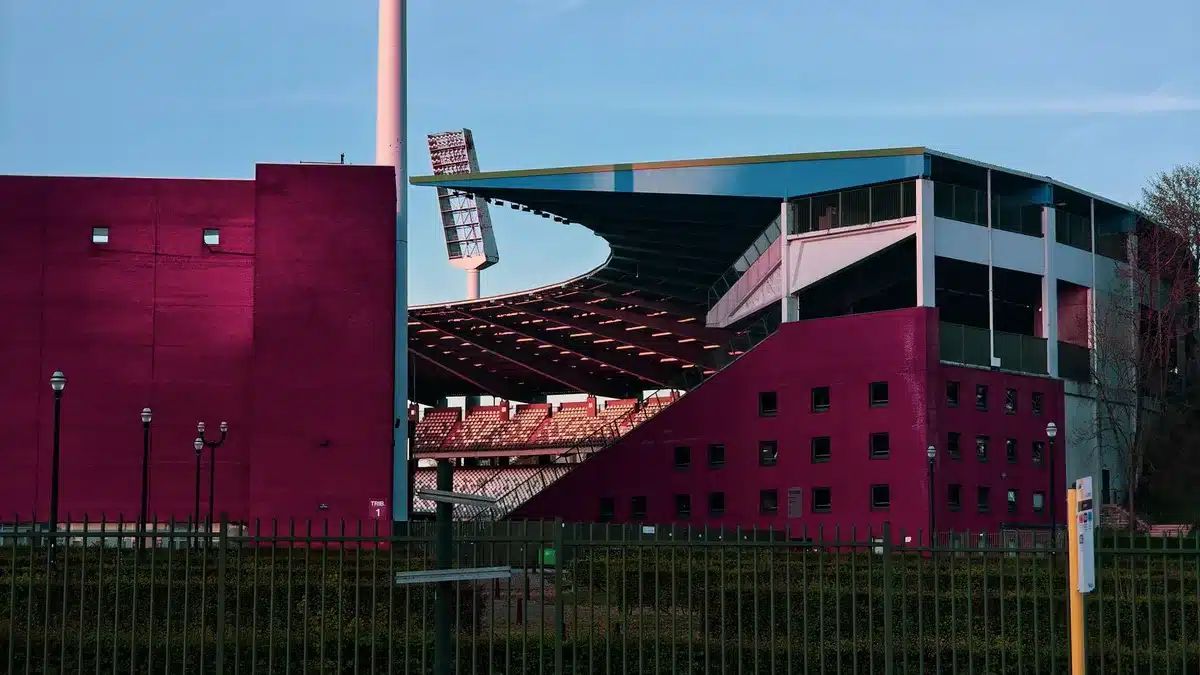 A packed stadium in the late afternoon light, rows of empty red seats gradually filling with spectators, floodlights beginning to glow, the pitch pristine and marked, anticipation palpable in the air