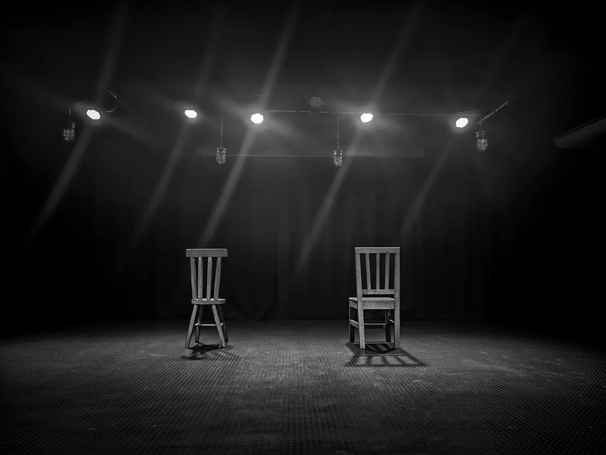 An empty theater stage with a single spotlight illuminating the wooden planks, a vintage microphone stand center stage, theatrical curtains framing the sides, soft shadows suggesting an era of classic drama.