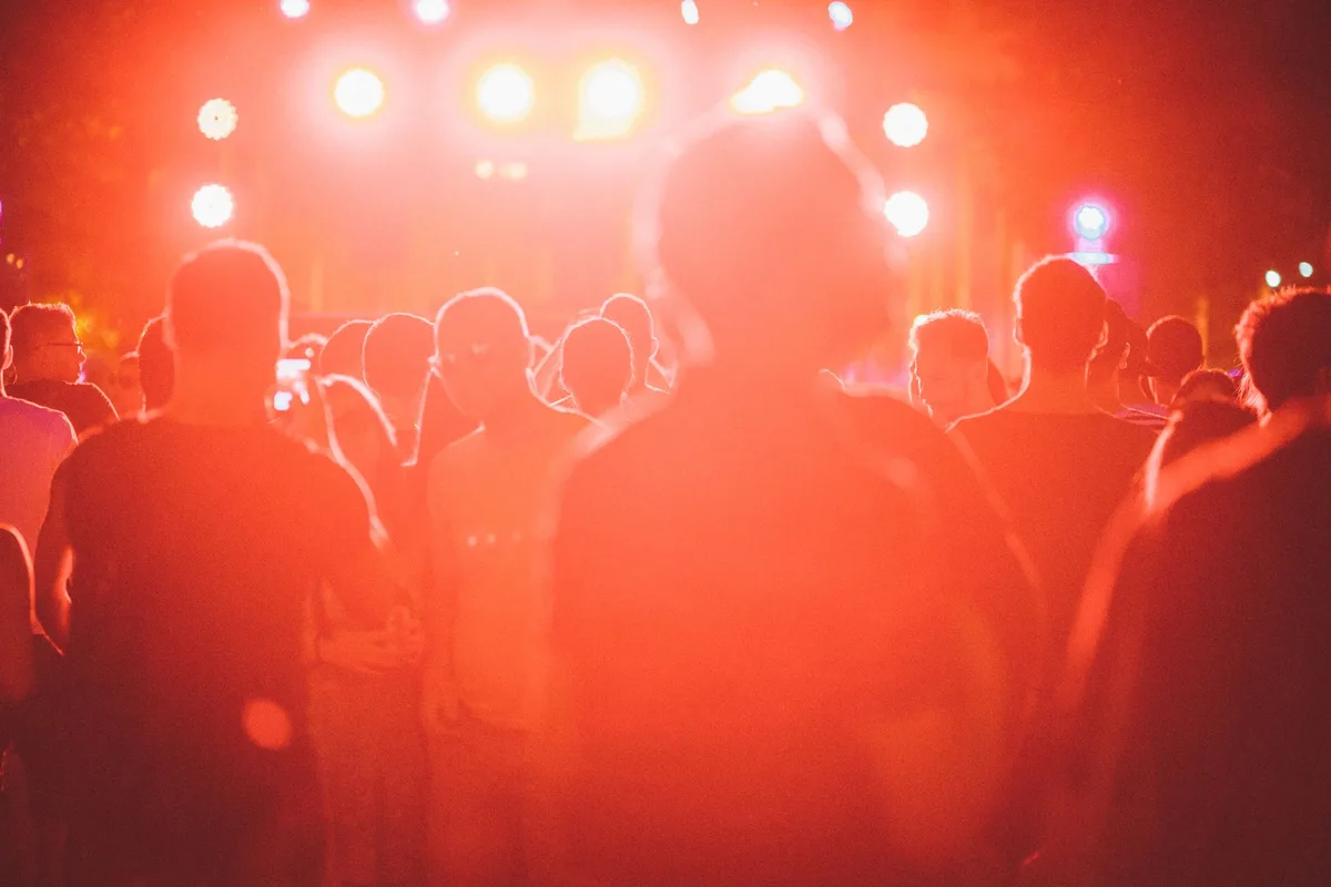 A stage under bright floodlights with a silhouetted crowd of anonymous fans with raised arms, swaying and dancing together, the energy of celebration visible in motion blur, confetti or mist suspended in the light beams