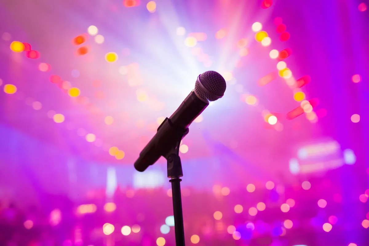 A close-up of a generic stage microphone under a single spotlight against a dark background, a coiled cable trailing into shadow, dust particles visible in the bright beam