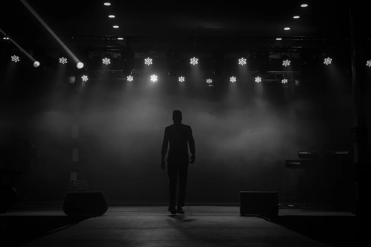 A stage with a single microphone stand under a spotlight, the venue empty and silent, a single abandoned guitar resting against an amplifier, concert lights dimmed in the background, the scene evoking an unperformed show