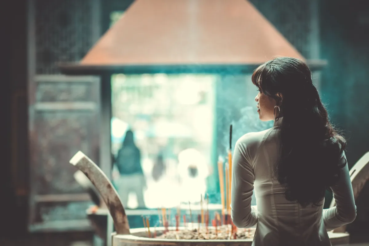Woman in a quiet moment of reflection, hands clasped together, soft natural light through a window, emotional but composed expression, intimate domestic setting suggesting solitude and contemplation