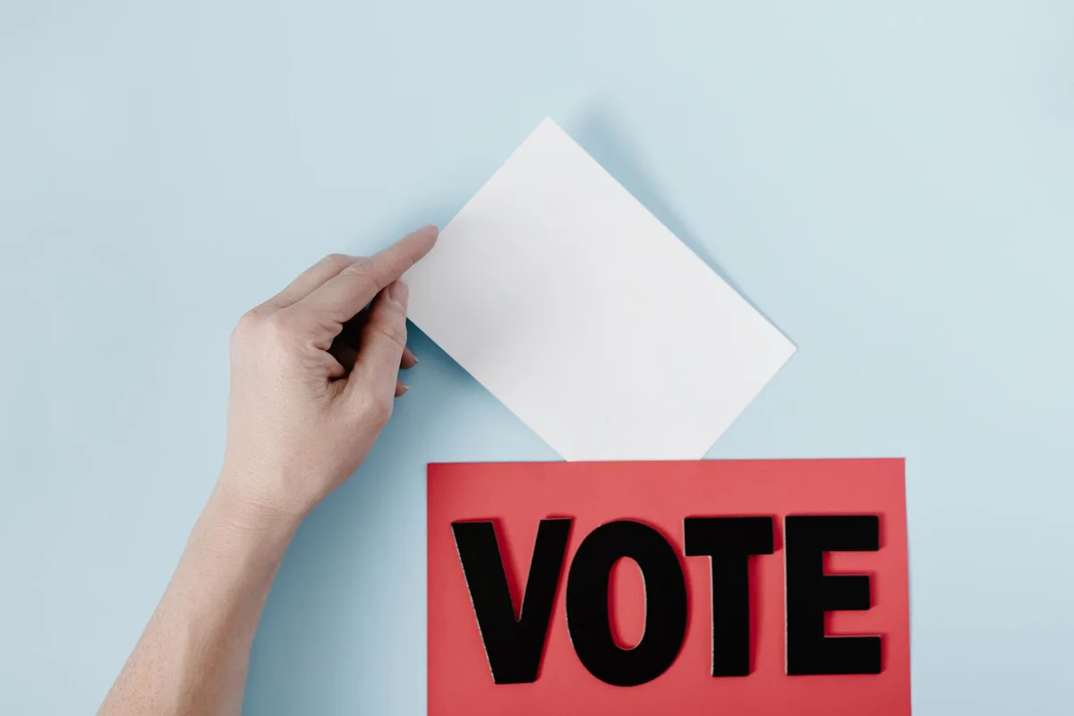 A ballot box and voting papers arranged on a desk with campaign materials in the background, representing the electoral process in a generic voting location