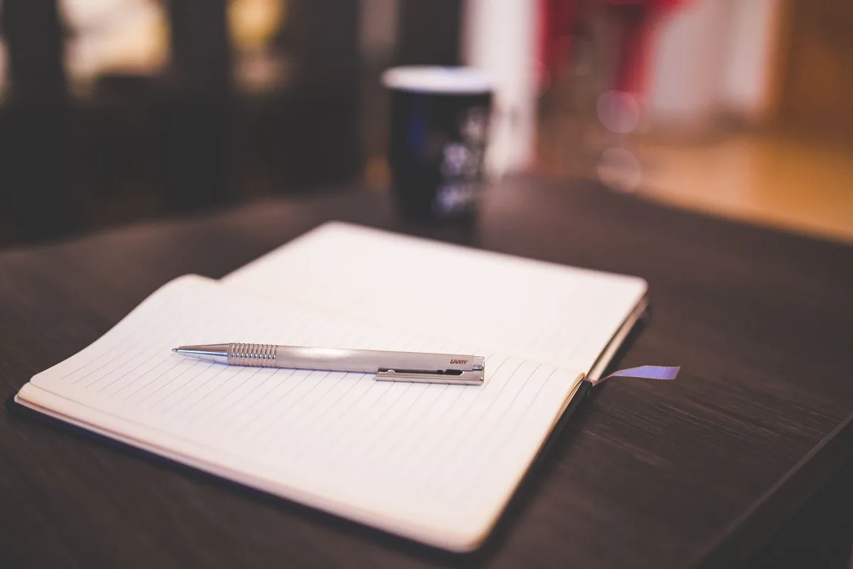A desk scattered with manuscript pages and a fountain pen, notebook open with handwritten notes, warm lamplight casting shadows, literary environment suggesting creative work