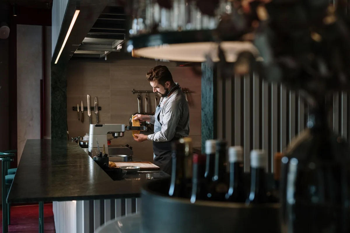 A white wine glass catching light on a professional kitchen counter, beside an empty plate, soft shadows across a stainless steel surface