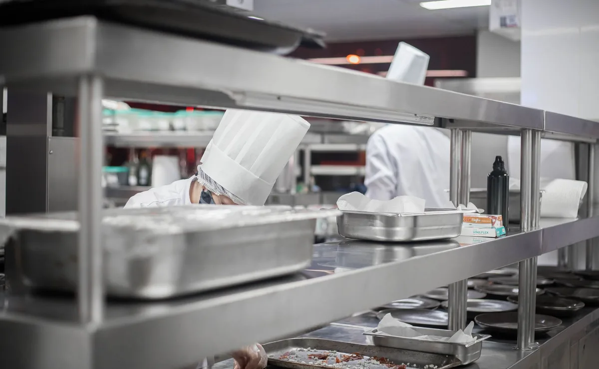 An empty kitchen station with a single apron hanging on a hook, soft overhead lighting creating shadows, a blurred backdrop of stainless steel equipment