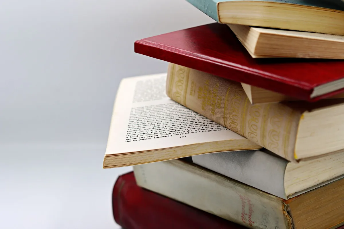 Stack of classic novels arranged on a wooden surface with warm overhead lighting, spines visible showing author names, suggesting intellectual exploration
