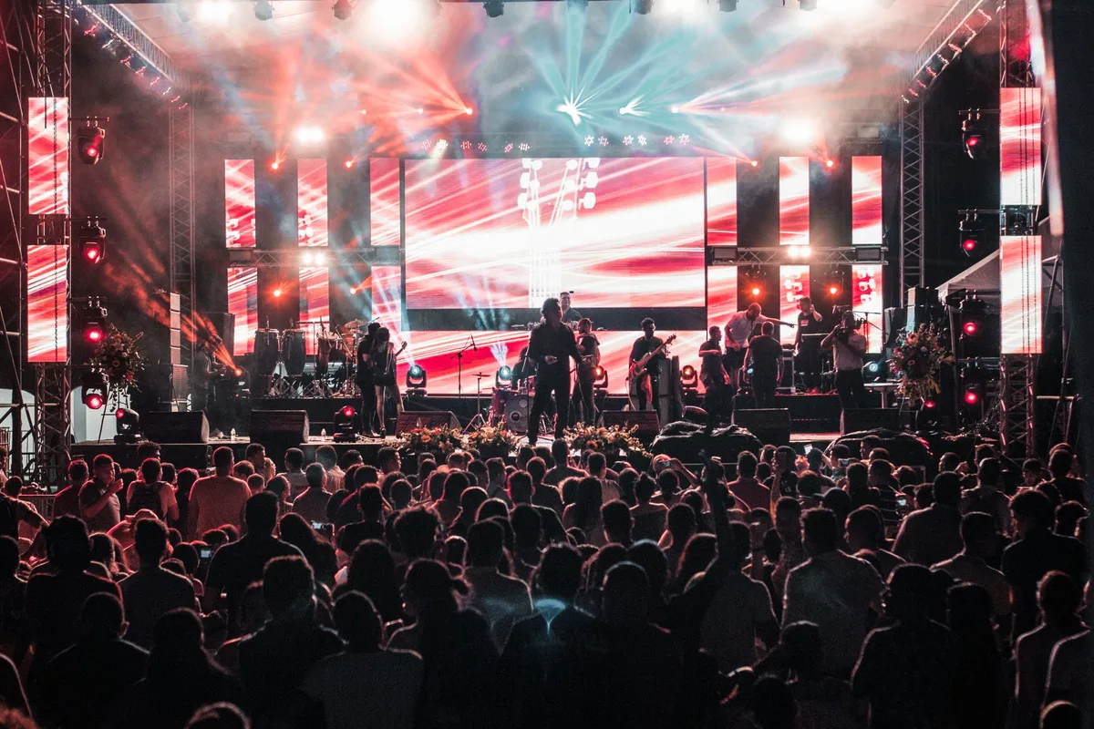 A stage spotlight on a microphone stand with silhouettes of a crowd with raised arms and phones recording, confetti suspended mid-air, warm lighting against dark background