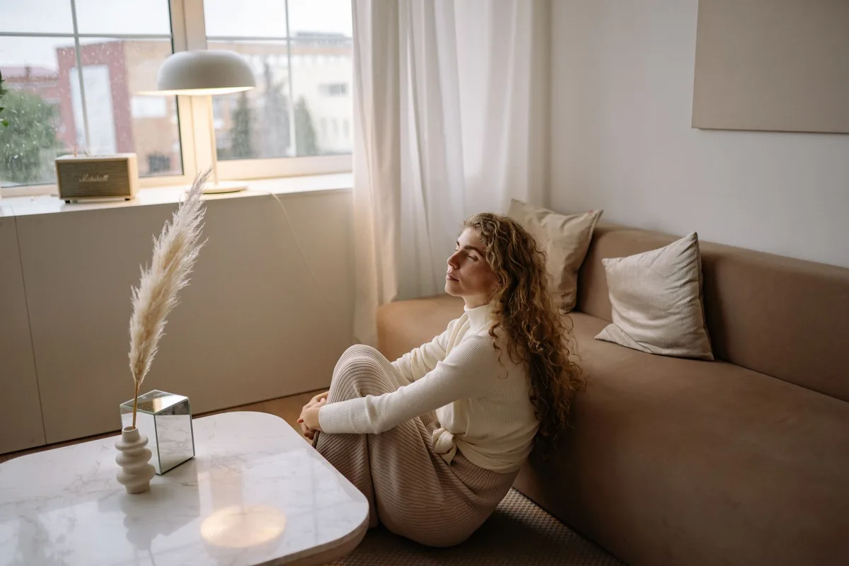 A woman sitting alone in a modern living room, hands clasped, looking thoughtful and serene, with soft natural light filtering through windows, conveying inner strength and peace after personal reflection