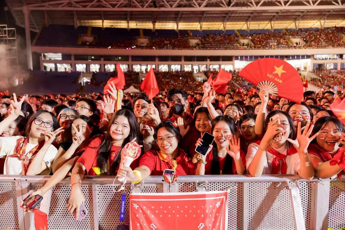 Anonymous crowd of fans under stadium floodlights, arms raised in celebration, silhouettes dancing and cheering, confetti or dust suspended in the light beams, faces blurred in motion.