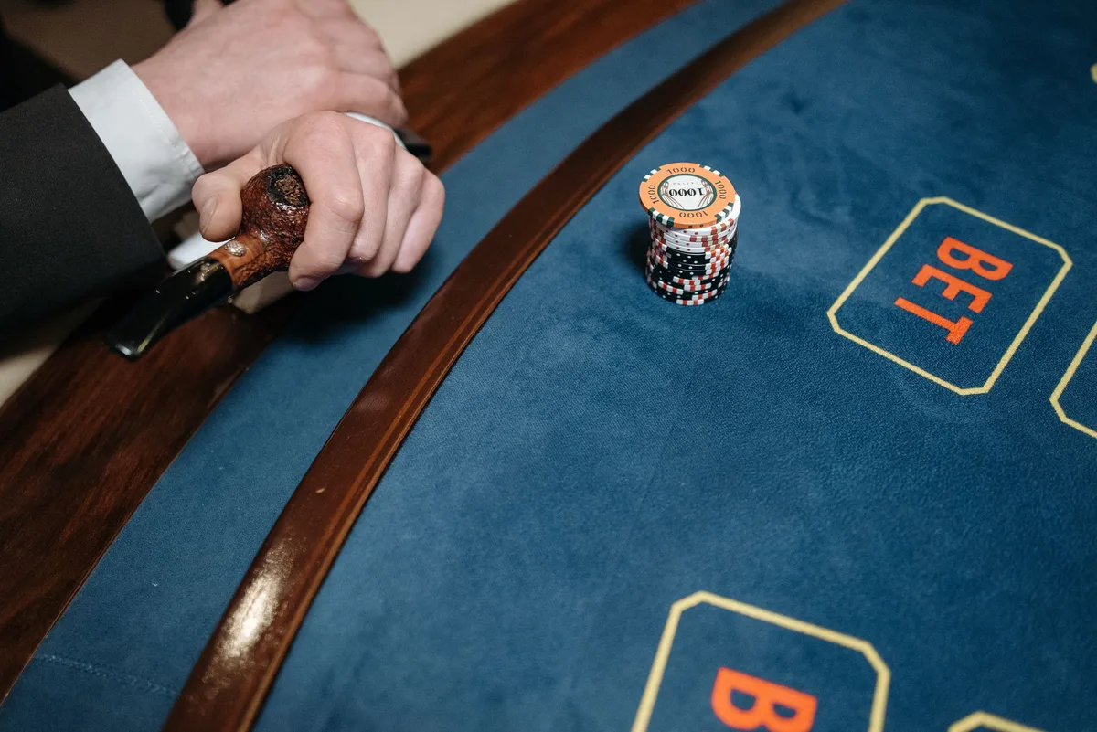 A shadowy figure at a gaming table with playing cards and poker chips scattered, dim lighting creating tension and desperation, anonymous hands reaching across the table