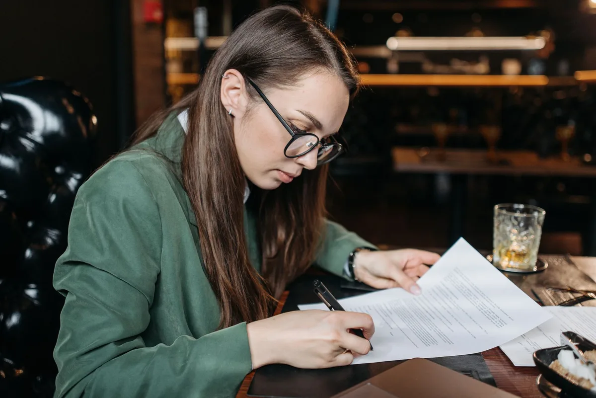 Anonymous woman in business attire examining documents at a desk with a pen, focused expression, professional office setting with soft lighting