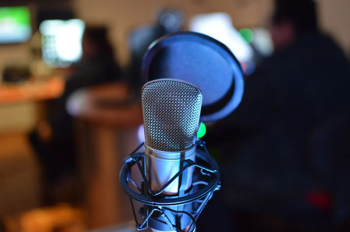 A radio broadcasting studio with microphones, mixing boards, and monitors displaying news feeds, modern equipment illuminated by studio lights, technicians in the background managing live transmission
