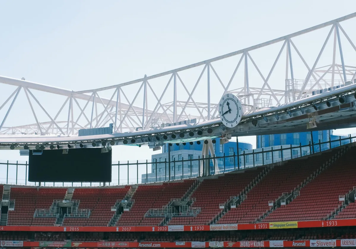 A broadcasting booth overlooking a football pitch, microphone and headphones on a desk, evening stadium lights in the background, focused on the technical setup.