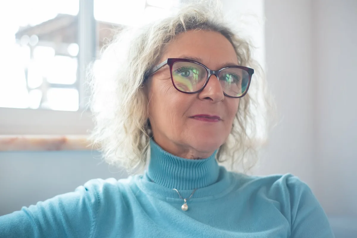 A woman in recovery wearing dark protective sunglasses indoors, sitting in a medical or home setting, with medical supplies visible on a side table, representing post-surgical care and healing.