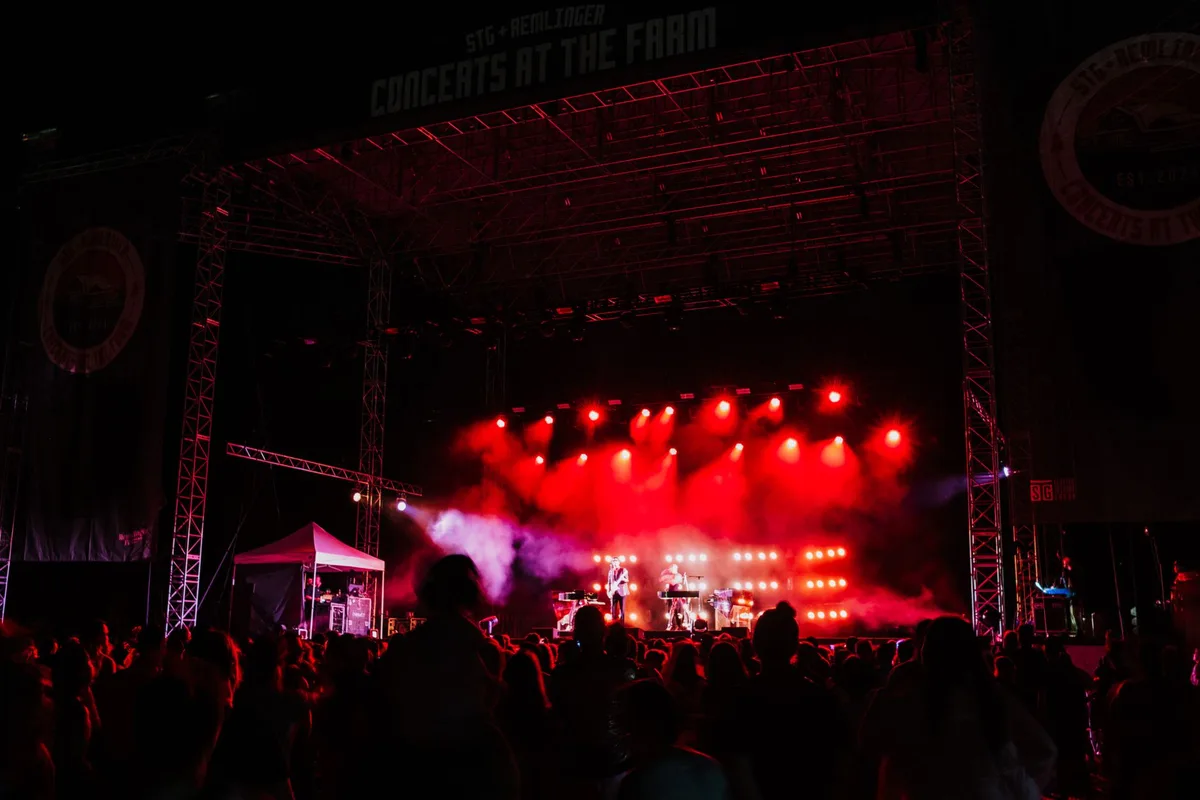 Vibrant festival stage with dramatic lighting, massive screens, and a roaring crowd of silhouettes with raised arms under night sky, confetti suspended in air