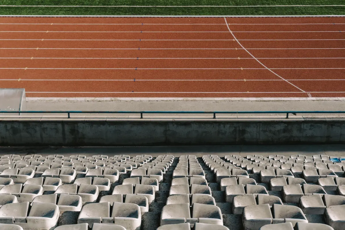 Empty soccer stadium seats at dusk, with one solitary spotlight illuminating the center field, shadows stretching across the pitch