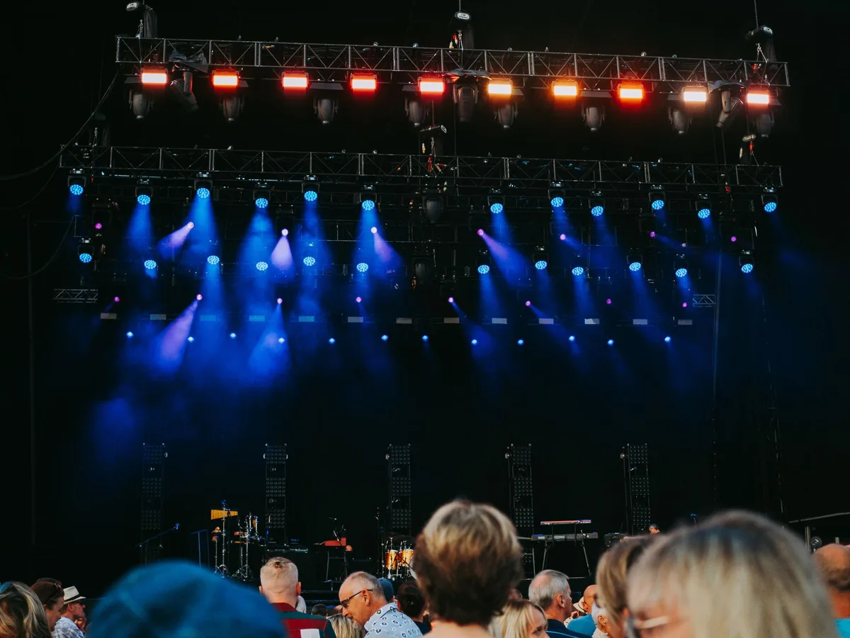 Festival stage at night with colorful lighting, crowds of anonymous spectators with raised arms and movement blur, urban historic architecture visible in background, collective energy and celebration