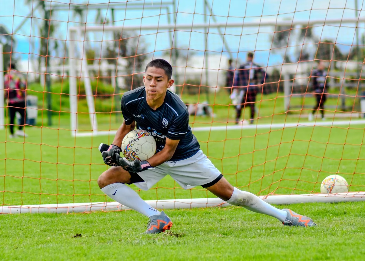A goalkeeper making a diving save during a Copa Libertadores match, hands extended toward the ball, stadium lights bright overhead, tension and focus visible in the moment