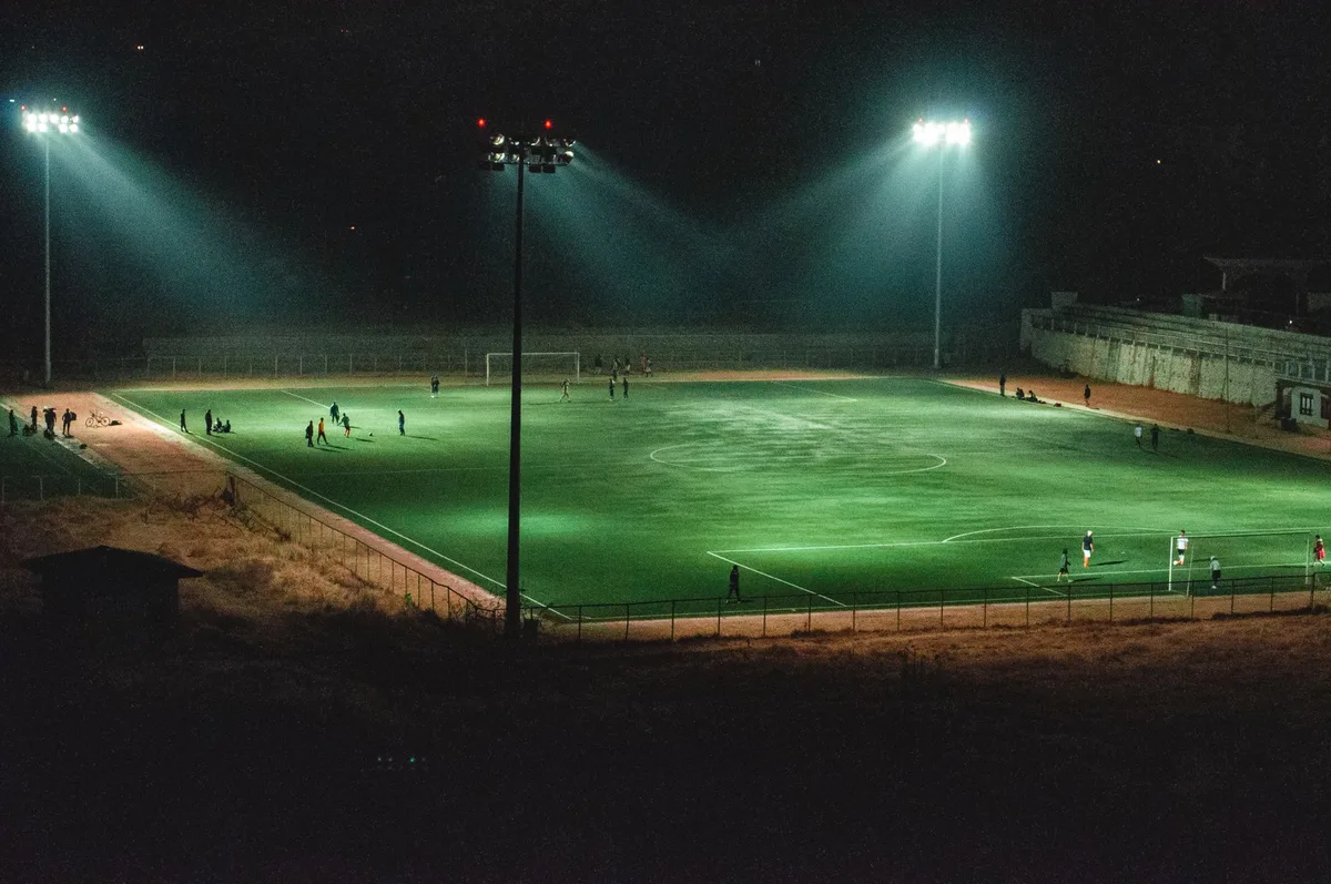 A football pitch at dusk with stadium lights beginning to illuminate, a single ball on the grass, empty benches in the background, cool blue and orange lighting tones