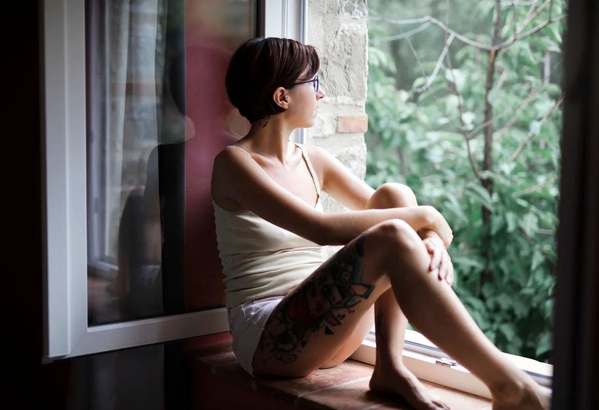 Silhouette of a woman looking out a window at dusk, hands pressed against glass, soft warm light behind her, solitude and reflection