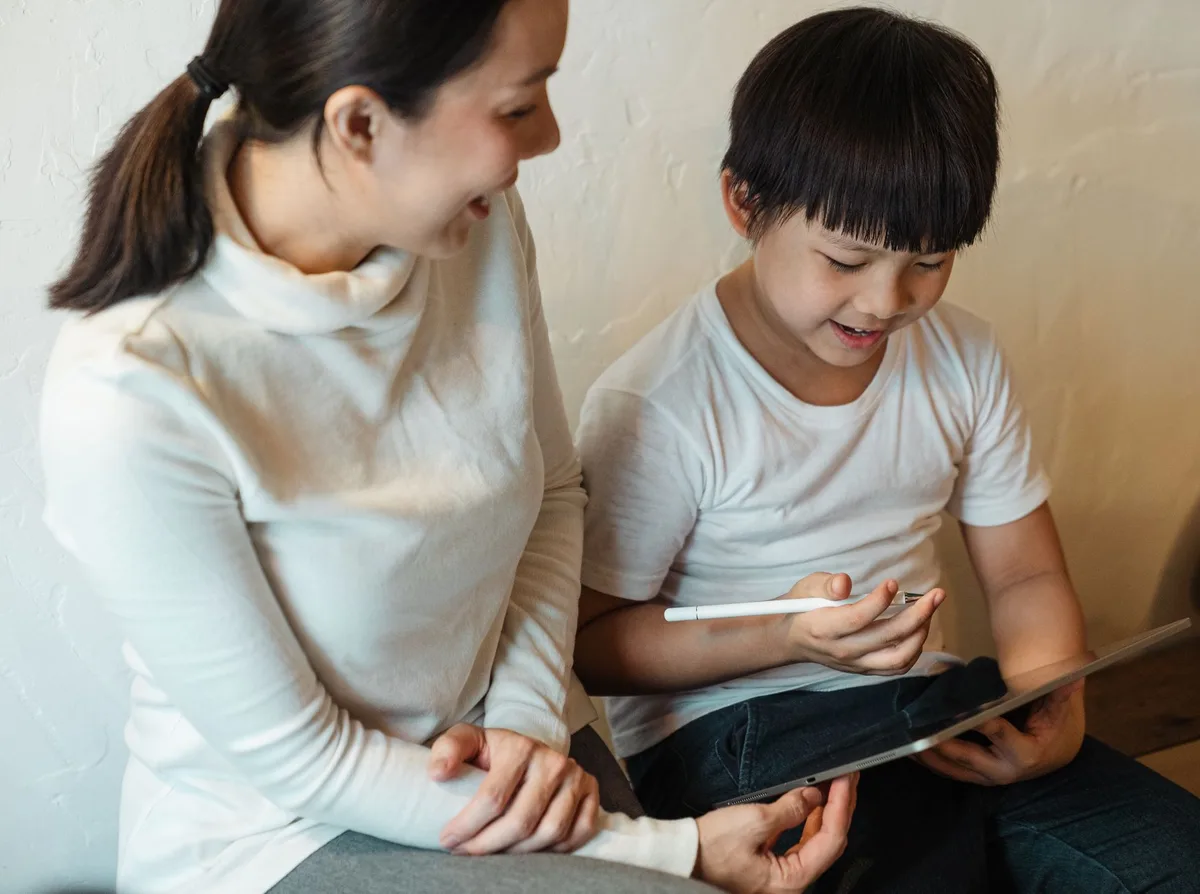 A mother and adult son sitting together in warm afternoon light, looking at old photographs spread on a table, genuine smiles and connection visible