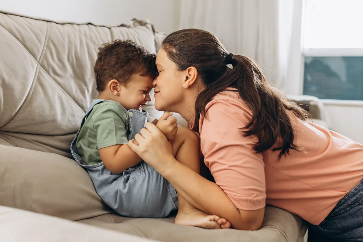 A mother and young child in a warm, domestic moment—hands holding, or a child reaching toward an adult figure—soft natural light filtering through, no faces visible, conveying unconditional bond and presence