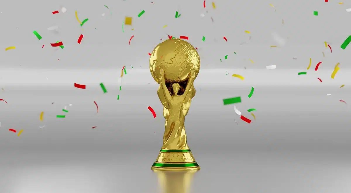 Jogador português em primeiro plano levantando os braços em celebração, com companheiros ao fundo em euforia, sob luzes do estádio ao anoitecer, confete caindo.