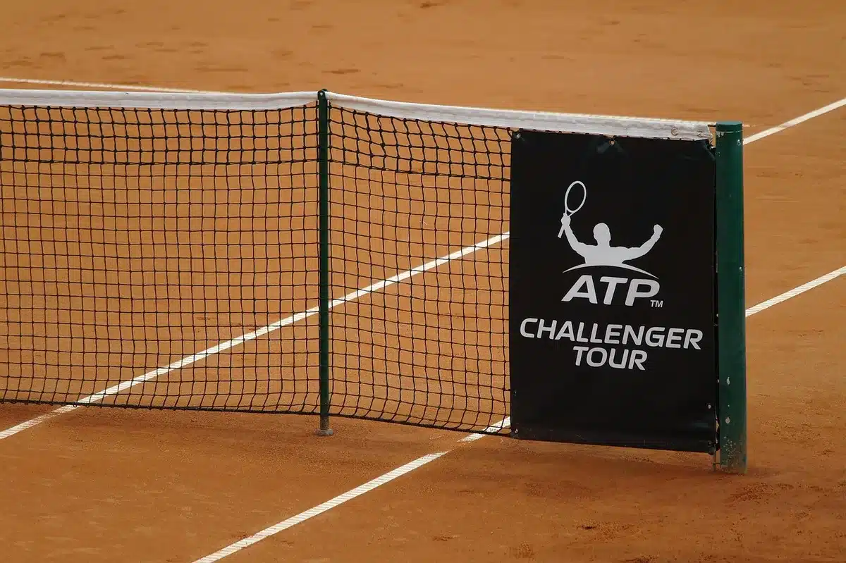 Close-up of an empty tennis court at night with a single spotlight on the net and a lone tennis ball near the baseline, empty stands blurred in the background