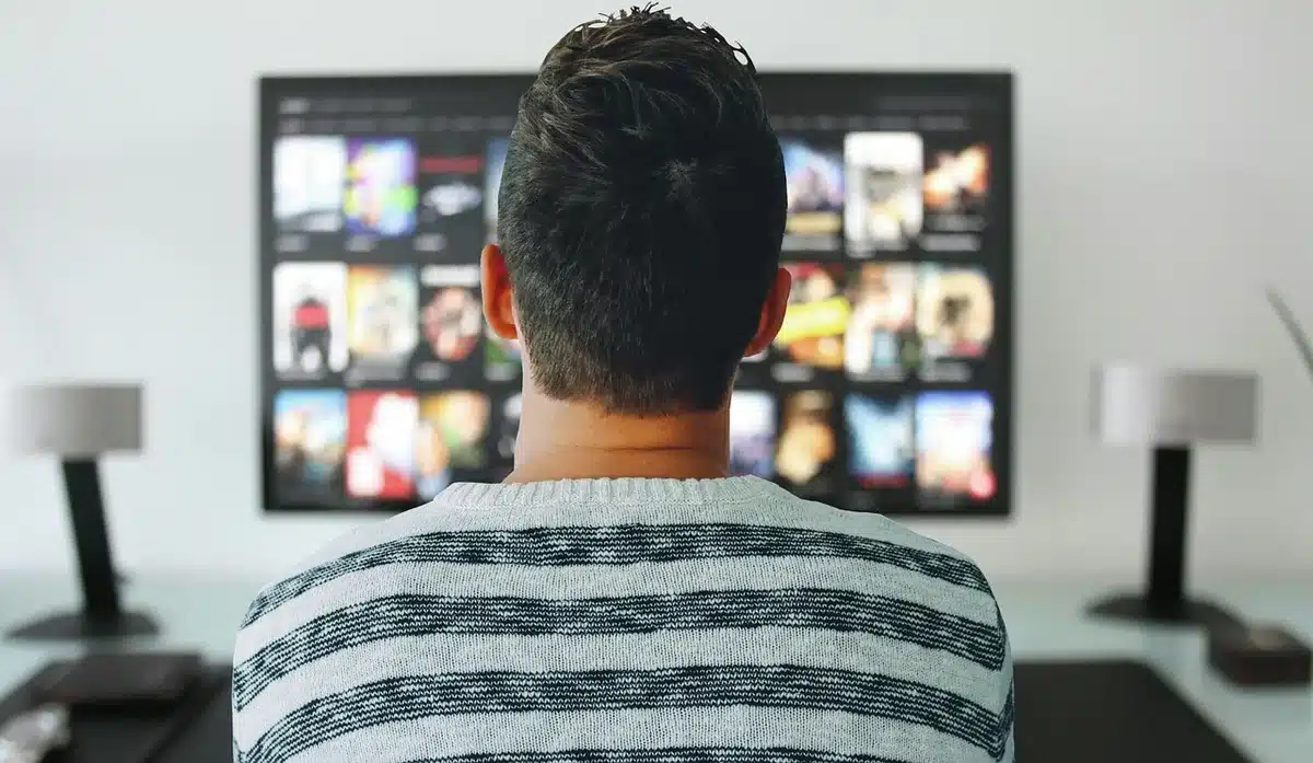 A television screen glowing in a dark room, displaying the opening frame of a comedy series, with warm light reflecting on the viewer's face in silhouette, conveying anticipation and engagement.